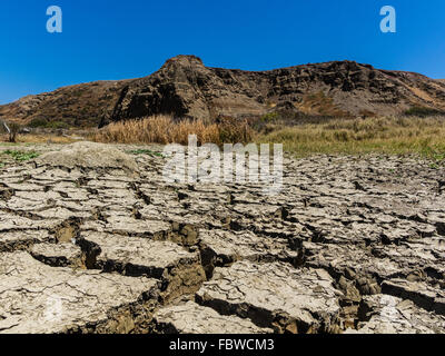 A close-up view of dried out mud flats with large cracks and peeling tops in between flat areas and hill in background. Stock Photo