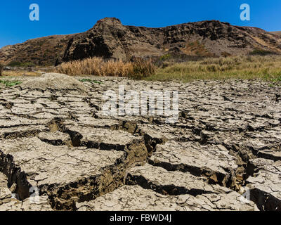 A close-up view of dried out mud flats with large cracks and peeling tops in between flat areas and hill in background. Stock Photo