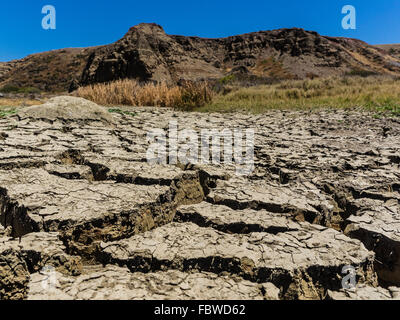 A close-up view of dried out mud flats with large cracks and peeling tops in between flat areas and hill in background. Stock Photo