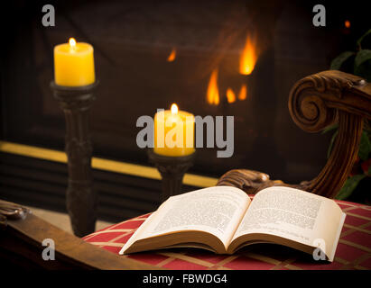 Paperback book open on chair by fire and candle Stock Photo