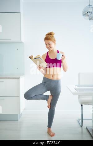 Young active woman with cup of tea reading book and doing yoga exercise in the kitchen Stock Photo