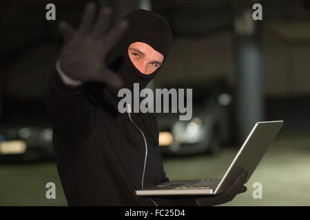 Hacker in balaclava gesturing and using laptop Stock Photo