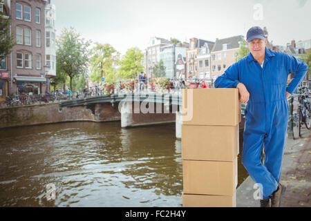 Composite image of happy delivery man leaning on pile of cardboard boxes Stock Photo