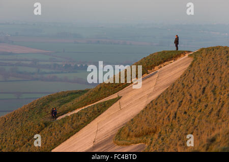 Bratton, Wiltshire, UK. 20th January 2016. The oldest chalk White Horse in Wiltshire was bathed in the orange glow of the setting sun this evening as temperatures began to plummet once more below zero. A couple attempt to slide down the giant head of the horse to take a selfie on its' nose. Credit:  Wayne Farrell/Alamy Live News Stock Photo