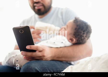 Father bottle feeding baby son and using cell phone Stock Photo