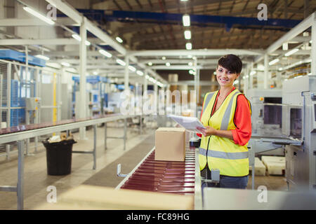 Portrait smiling worker checking cardboard boxes on conveyor belt production line in factory Stock Photo
