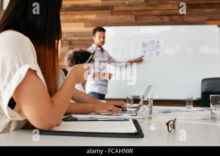 Closeup image of young woman sitting in a meeting in boardroom with man giving presentation in background. Focus on hands of fem Stock Photo