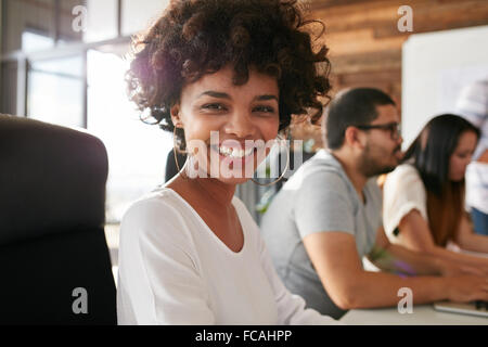 Closeup portrait of cheerful young african woman sitting in conference room with coworkers in background. Creative team meeting Stock Photo