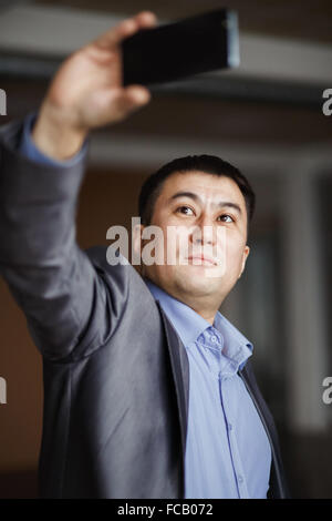 Businessman holding mobile smart phone using app doing selfie, preparing video call, wearing jacket. Selfie style photo, serious man in suit making self portrait for social networks. Stock Photo