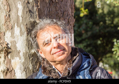 Middle-aged Caucasian man with gray hair dressed in black down jacket leaning against a tree trunk Stock Photo