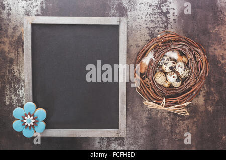 Easter background. Quail eggs in a nest, blackboard and Easter traditional gingerbread cookie in flower shape. Stock Photo