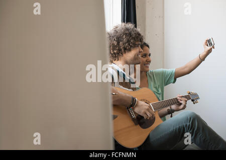 Loft living. A young man playing guitar and a woman beside him taking a selfy with a smart phone. Stock Photo
