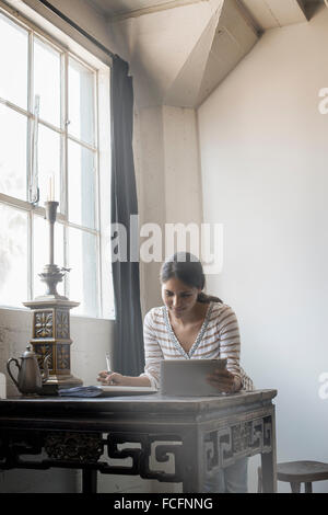 Loft living. A woman sitting at a table by a window using a digital tablet. Stock Photo