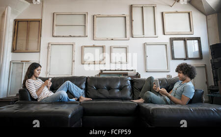 A young couple sitting on the sofa, one using a smart phone and one holding a digital tablet. Stock Photo