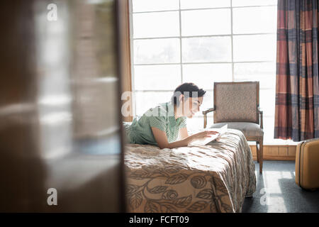 A young woman lying on a bed in a motel room using a digital tablet touchscreen checked curtains, mobile communications internet Stock Photo