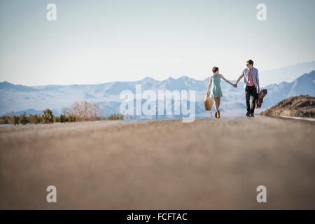 A young couple, man and woman walking hand in hand on a tarmac road in the desert carrying cases. Stock Photo