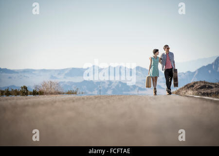 A young couple, man and woman, on a tarmac road in the desert carrying cases. Stock Photo