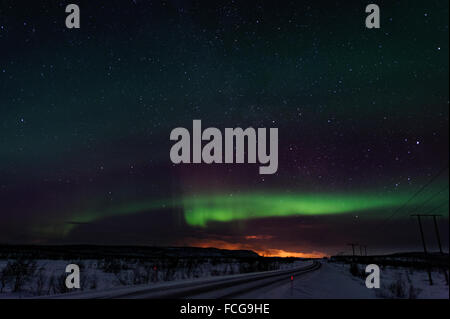 Northern lights and stars above a road with snow. The clouds on the horizon are illumintaed by the city lights of Alta, Norway Stock Photo