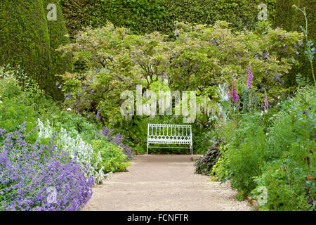 White metal bench under white fragrant wisteria tree at the end of stone pathway in summer garden with cottage flowers in bloom
