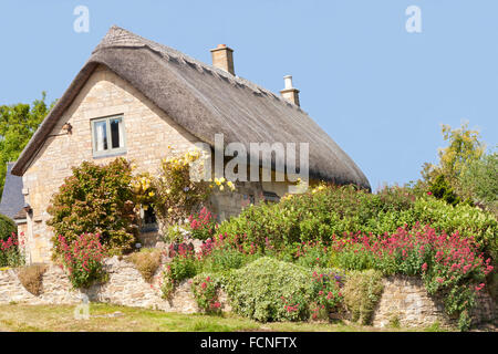 Traditional old thatched roof stone Cotswold cottage holiday home with roses on the wall and flowers in the summer garden Stock Photo