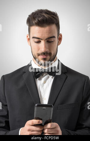 Young man in tuxedo with bow tie holding mobile phone texting message. Portrait over gray studio background. Stock Photo
