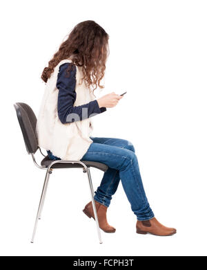 back view of woman sitting on chair and looks at the screen of the tablet. Rear view people collection. backside view of person. Isolated over white background. A girl in a white tank top sitting on a chair looking at the phone Stock Photo