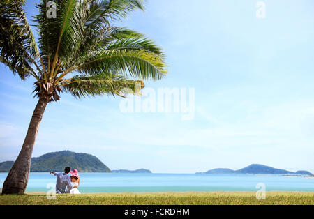 Happy young caucasian couple sitting under palm near the sea Stock Photo