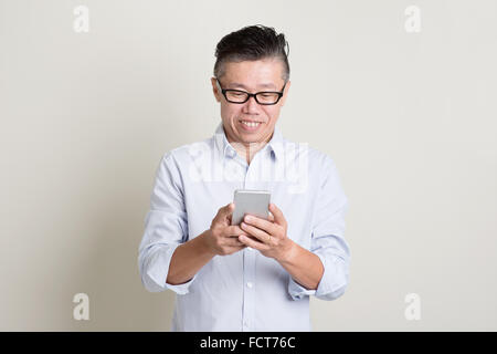 Portrait of single mature 50s Asian man in casual business using mobile apps on smartphone and smiling, standing over plain back Stock Photo