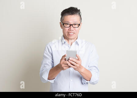 Portrait of single mature 50s Asian man in casual business using mobile applications on smartphone and smiling, standing over pl Stock Photo