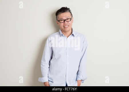 Portrait of happy single mature 50s Asian man in casual business smiling, standing over plain background with shadow. Stock Photo