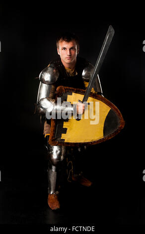 Studio shot of young man dressed as medieval knight armed with sword and shield in ready-to-fight position over black background Stock Photo