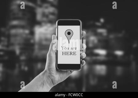 Female hand holding a mobile phone vertically with "you are here" text and placemark on the screen. On blurred urban background Stock Photo