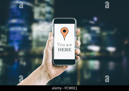 Female hand holding a mobile phone vertically with "you are here" text and placemark on the screen. On blurred urban background Stock Photo