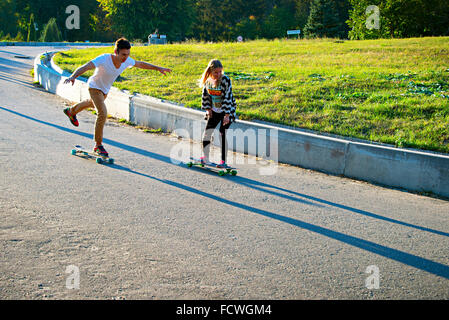 Young couple riding the longboards down the hill at sunset in a park. Stock Photo