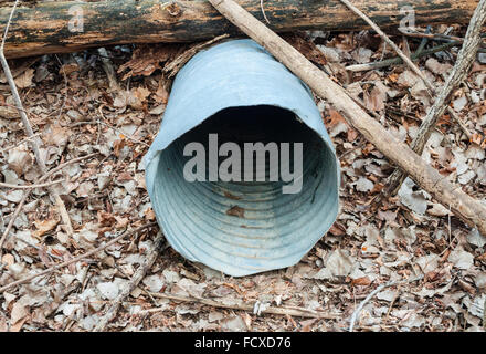 Corrugated empty metal drain pipe under log and branches, on ground covered in dead leaves. Stock Photo