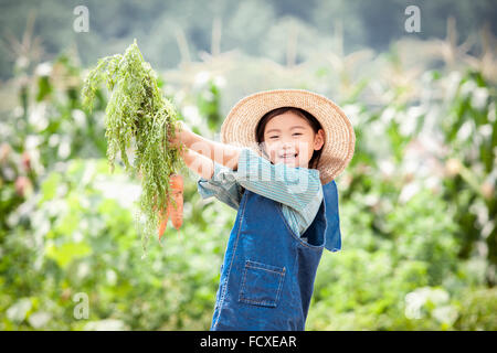 Girl in a straw hat holding carrots up and smiling at field Stock Photo