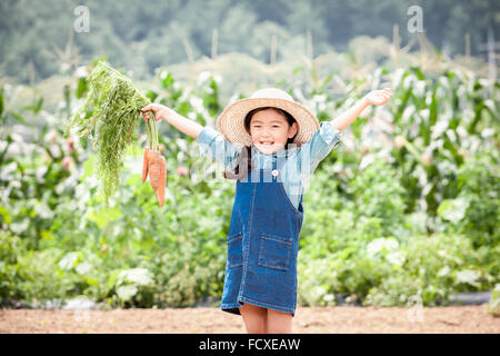 Girl in a straw hat holding carrots up and smiling at field Stock Photo