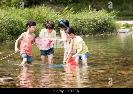 Four kids having fun for summer vacation in the water of the stream holding a scooping net for fishing and a bucket Stock Photo
