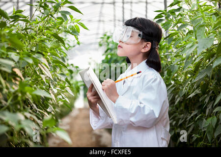 Girl in white gown and laboratory goggles writing down on a notebook while looking at the plants in green house Stock Photo