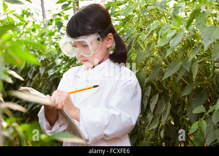 Girl in white gown and laboratory goggles writing down on a notebook surrounded by plants Stock Photo