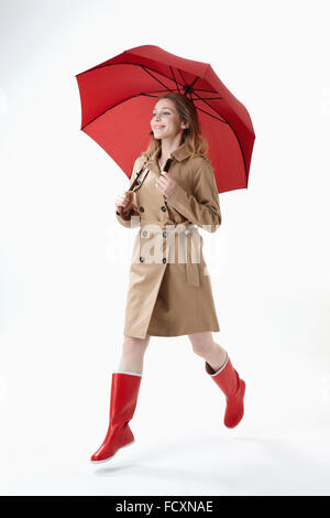 Young smiling woman wearing red umbrella, trenchcoat and red boots running Stock Photo