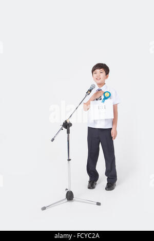 Elementary school boy in school uniform standing behind a standing microphone and wearing a numbering tag and a badge for Stock Photo
