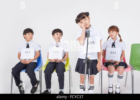 Elementary school girl standing with a microphone in worried face and three other students sitting behind her looking at her Stock Photo