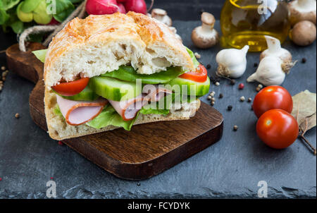 Rustic sandwich with ham, ciabatta bread,fresh salad,tomatoes and cucumber on wooden cutting board Stock Photo