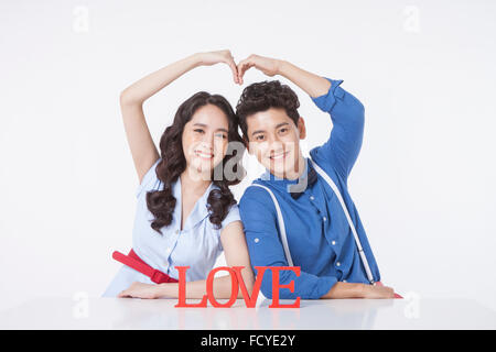 Couple in retro style making a heart shape with their hands up seated at desk behind solid word LOVE in red standing at desk Stock Photo