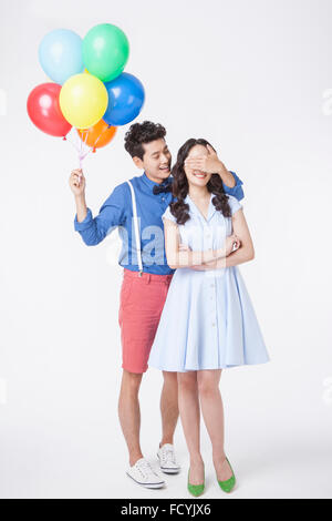 Man with colorful balloons in his hand covering woman's eyes with his hand behind her both in retro style outfits and standing Stock Photo