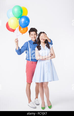 Man with colorful balloons in his hand covering woman's eyes with his hand behind her both in retro style outfits and standing Stock Photo