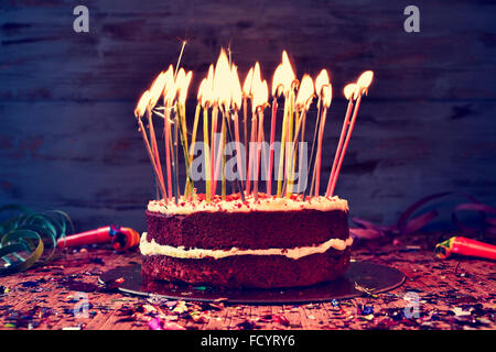 a cake topped with some lit candles before blowing out the cake, on a rustic wooden table full of confetti, party horns and stre Stock Photo