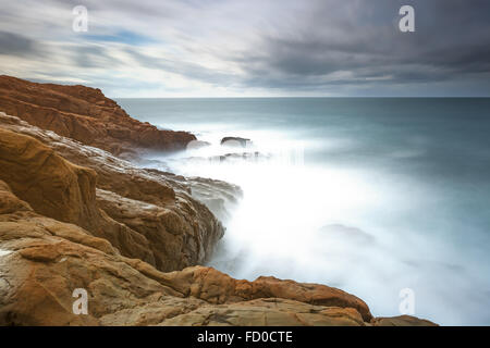 Dark red rocks, waves and foam, ocean under cloudy sky in a bad weather. Long exposure photography. Livorno, Tuscany, Italy Stock Photo