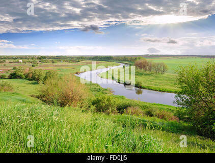Green meadow, village and river against a cloudy sky. Stock Photo